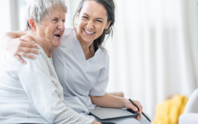 A smiling caregiver sits beside an older woman with her arm around her in a bright home setting.
