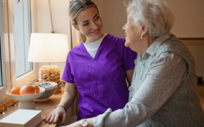 A caregiver in purple scrubs smiles while assisting an older woman as she stands beside a table in her home.