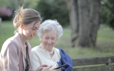 A caregiver and an older woman sit together on a park bench, smiling as they look at a smartphone.